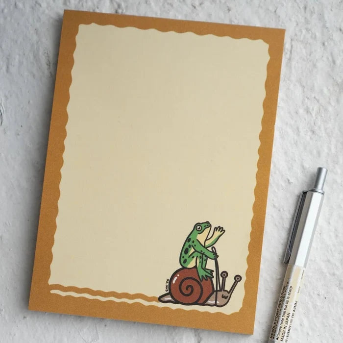 Stay Home Club Frog & Snail Ride Notepad featuring a cute frog riding a snail, brown border, and nature-inspired stationery design.