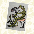 Forager Provisions LTD woven patch featuring a green frog sitting on a mushroom and holding a bouquet of flowers.