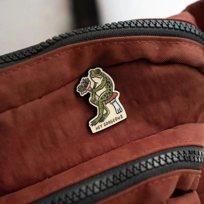 Stay Home Club Hey Gorgeous Pin decorates a brown bag near the zipper—chic positive accessory, uplifting motivational enamel pin.
