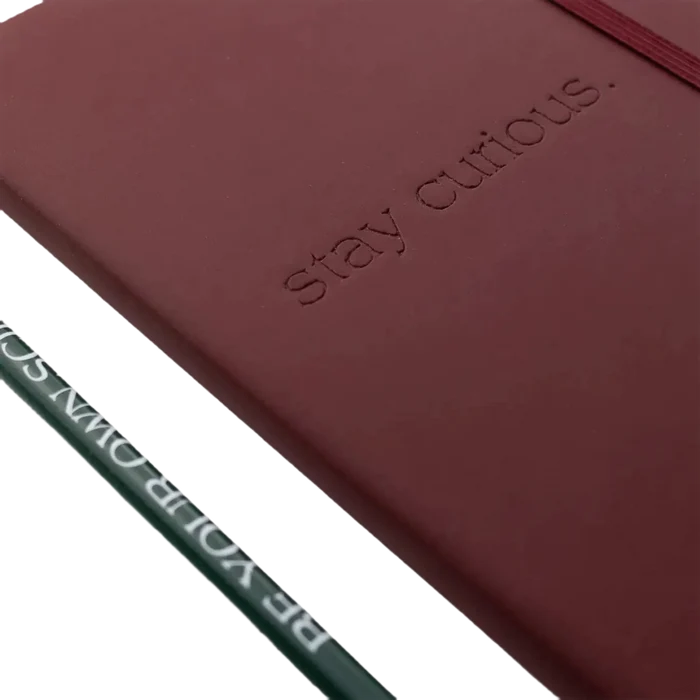 The Stemcell Science Shop's "Stay Curious Journal & Pencil Set," featuring a maroon, hardcover notebook with "stay curious" embossed on the cover—ideal for an observation journal—sits next to a green pencil inscribed with "BE YOUR OWN SCARS.