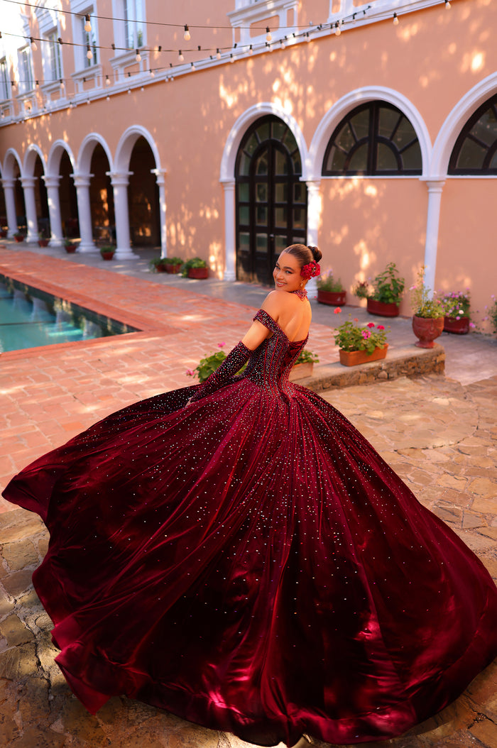 A woman in a sparkling burgundy ball gown poses by a pool near arched columned buildings.