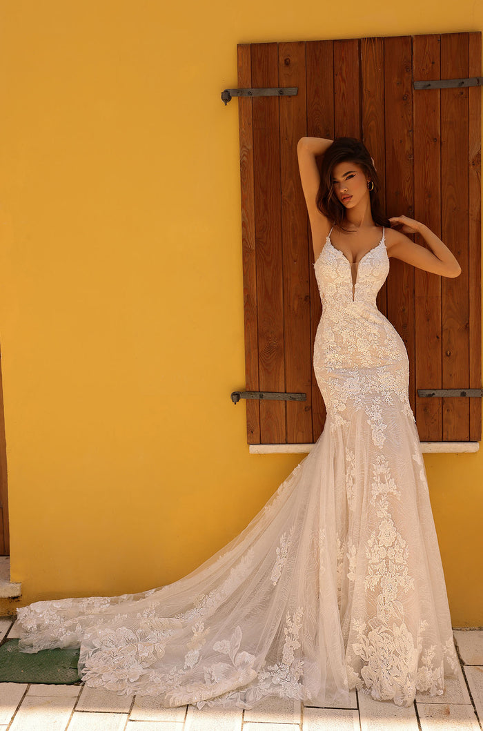 A woman in an intricate lace mermaid wedding dress poses against a yellow wall with wooden shutters.