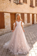 A bride in an off-shoulder lace wedding gown stands on cobblestone steps against a stone building backdrop.