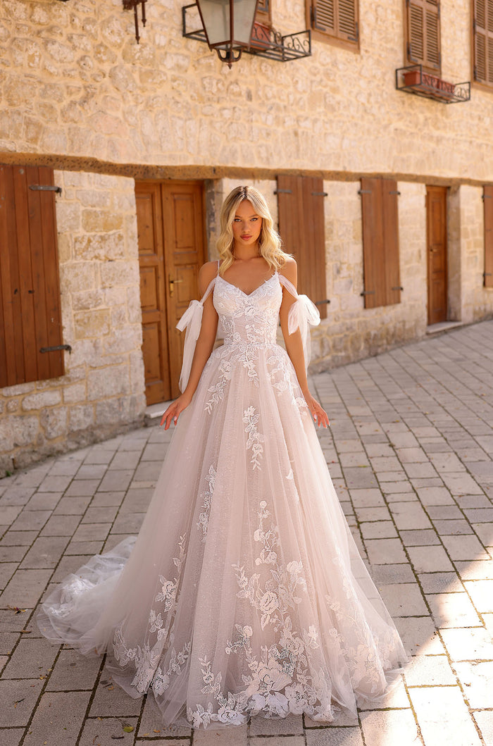 A bride in an off-shoulder lace wedding gown stands on cobblestone steps against a stone building backdrop.