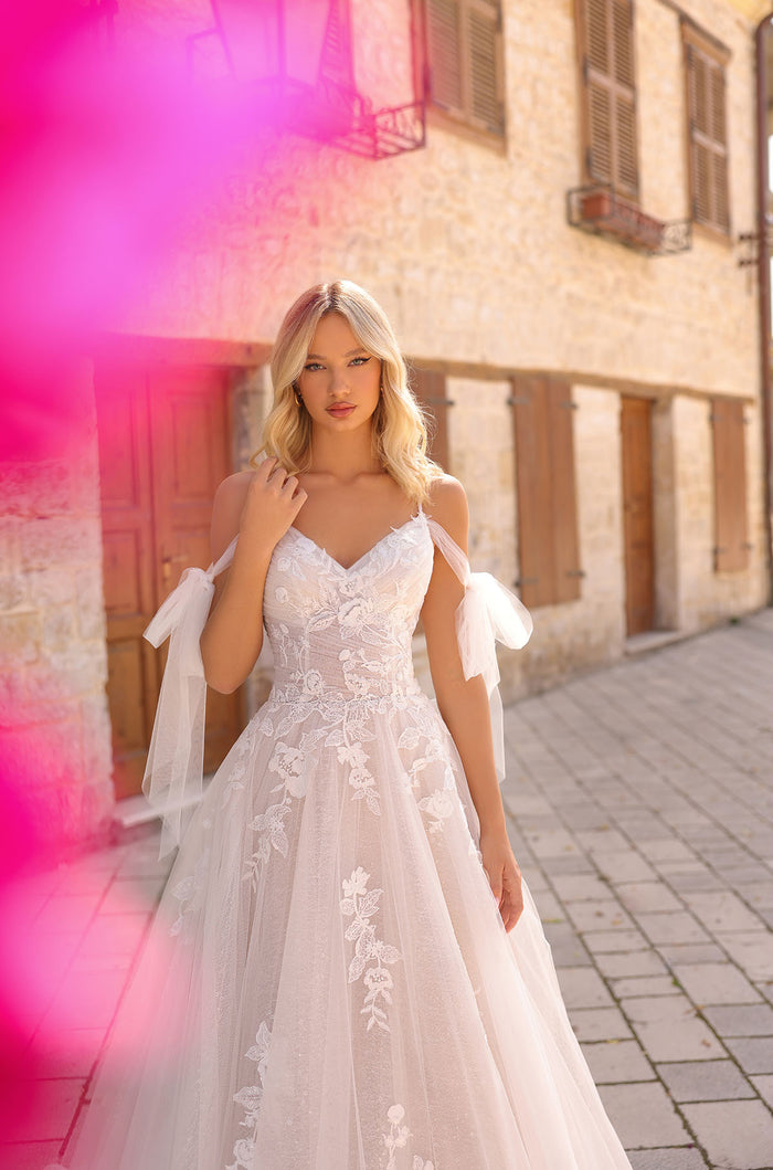 A blonde woman wears a white lace A-line wedding dress with off-shoulder straps on a sunlit stone street.