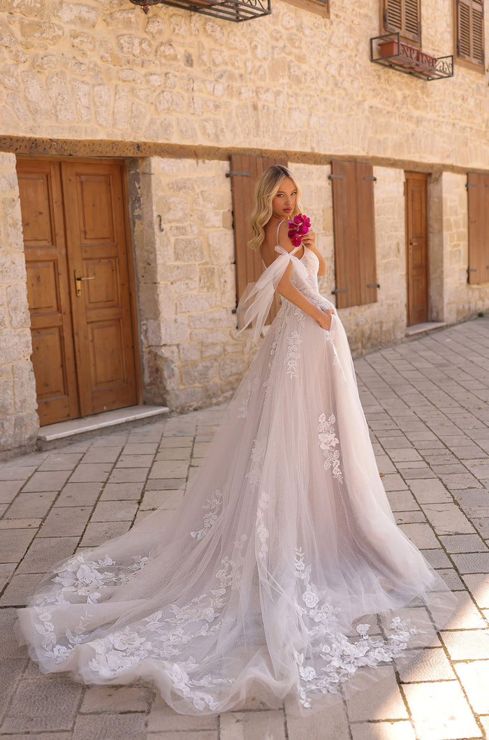 A bride in an off-shoulder lace wedding gown with a long train stands holding pink flowers against a stone building.