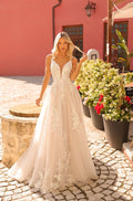 A blonde bride in an intricate lace A-line wedding gown stands on cobblestone beside flowering plants.
