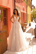 A slender model wears an intricate lace A-line wedding dress while standing on a sunny Mediterranean street.