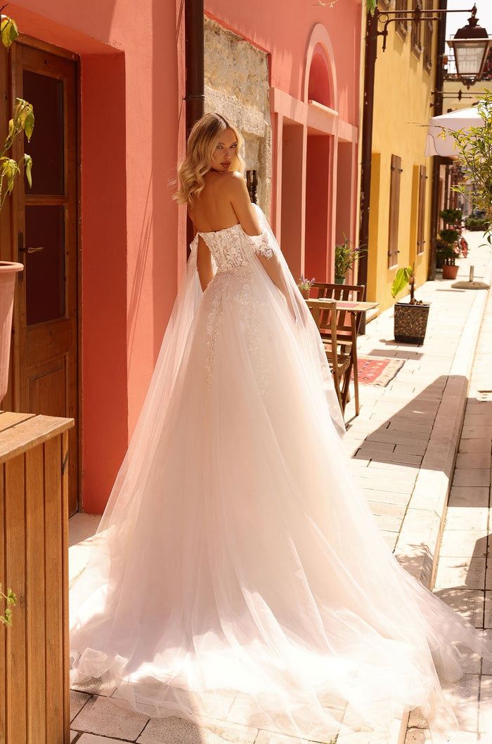 A bride in an elaborate white lace ball gown with a long train stands in a charming Mediterranean-style street.