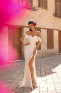 A woman in a white off-shoulder gown with a high slit poses elegantly on a stone-paved street.