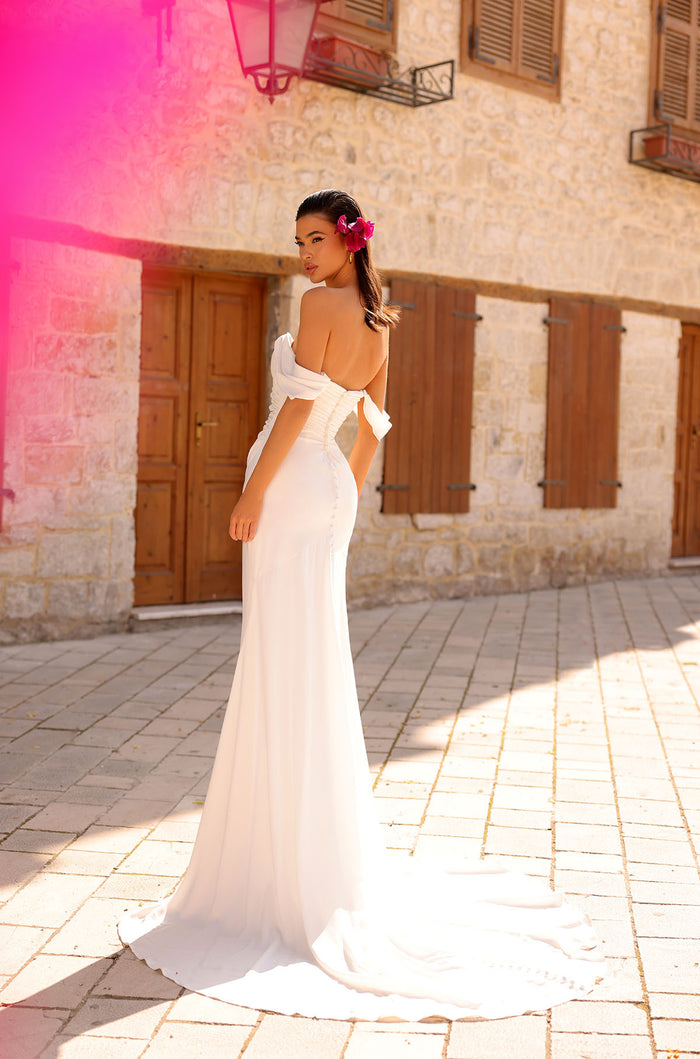 A bride in a white off-shoulder mermaid gown stands elegantly on a stone-paved street with pink-tinted lighting.