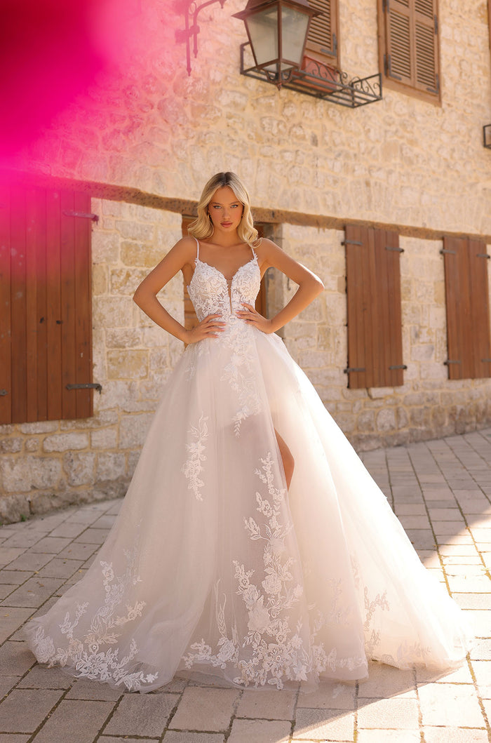 A bride in an elegant white lace ball gown stands confidently against a stone wall with wooden shutters.