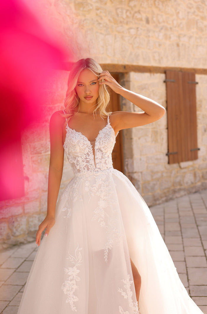 A blonde woman wears an elegant white lace wedding gown with a full skirt, standing before a stone wall.