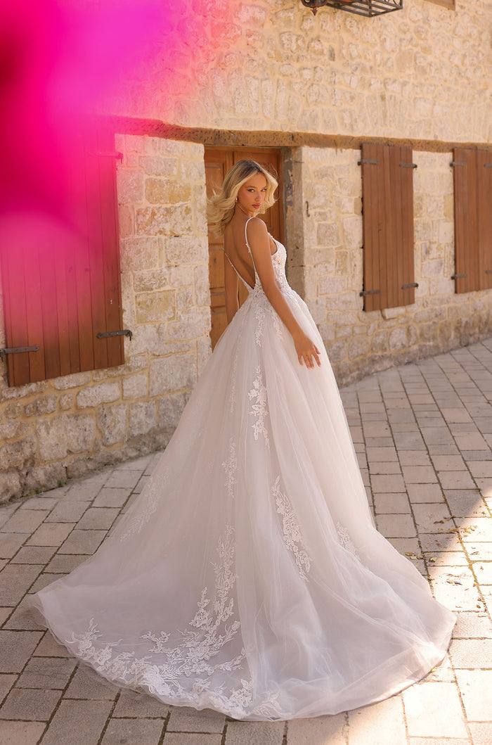 A bride in a white lace ballgown with an open back poses by a stone wall with wooden shutters.