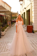 Blonde woman wears off-shoulder lace wedding gown with flowing tulle skirt in European street setting.