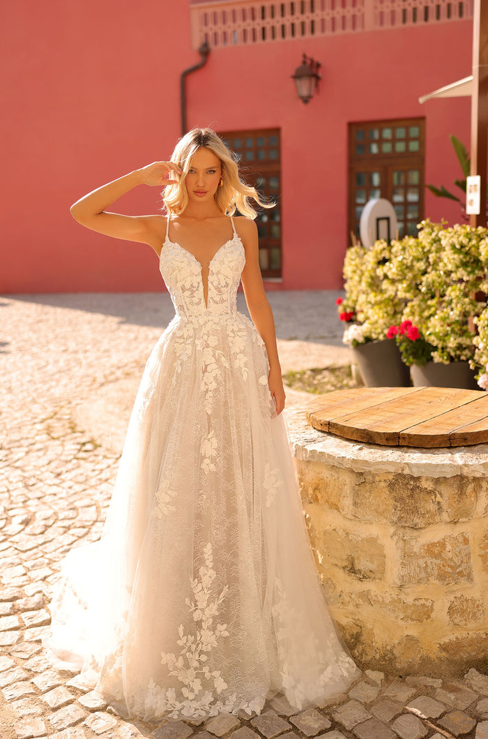 A blonde woman wears an intricate white lace wedding gown with floral details, standing on cobblestone near a pink building.