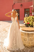 A blonde woman wears an intricate white lace wedding gown with floral details, standing on cobblestone near a pink building.