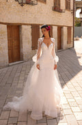 A bride in a white lace wedding gown with billowing sleeves stands on a stone-paved street near old buildings.