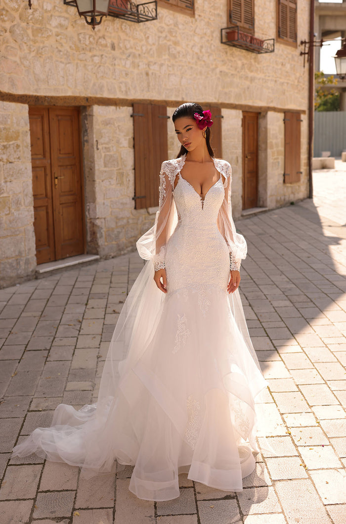 A bride in a white lace wedding gown with billowing sleeves stands on a stone-paved street near old buildings.