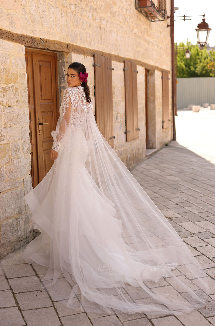 A bride in a white lace wedding dress with long train stands by a stone wall, wearing a pink flower in her hair.