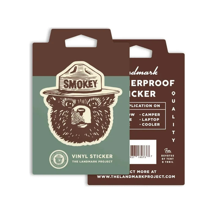The Landmark Project Smokey Bear Logo Sticker set includes two weatherproof vinyl stickers featuring Smokey Bear in a park ranger hat, with one sticker labeled "Smokey." These stickers are ideal for adventure cataloging or enhancing your collection of park memorabilia. The packaging emphasizes their waterproof quality.
