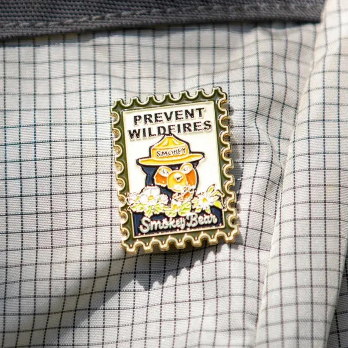 Forager Provisions LTD Smokey Bear enamel pin featuring a vintage postage stamp design on a grey checkered fabric.