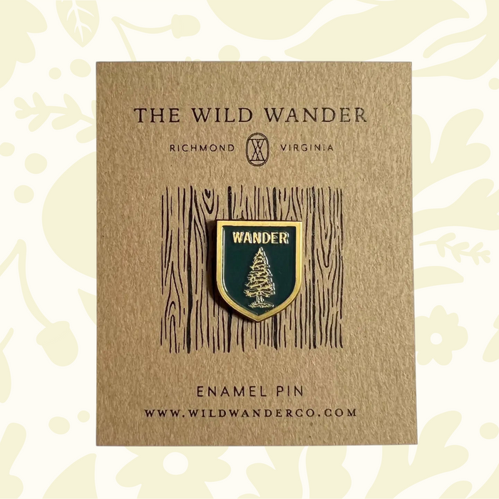 Green enamel pin with fir tree, WANDER text, on The Wild Wanders branded card—forest nature hiking accessory.