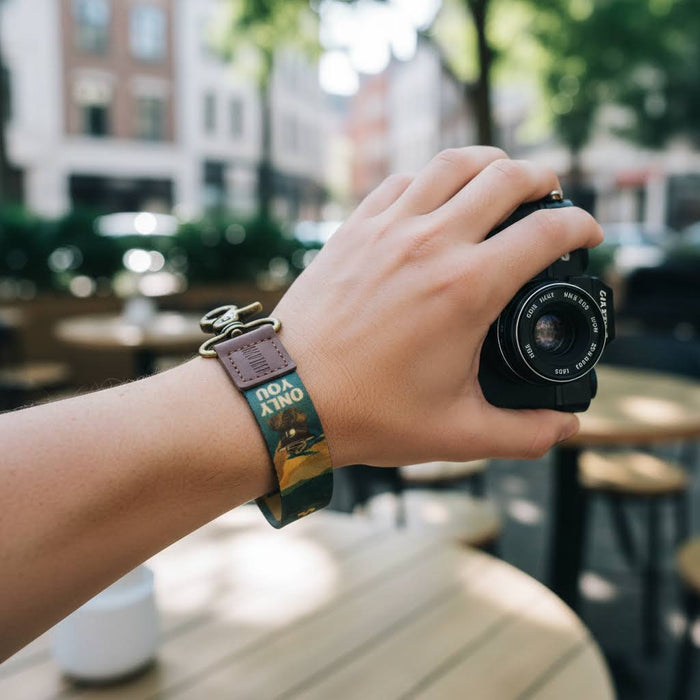 Hand holds camera with Wildtree Smokey Bear Wrist Keychain outdoors, raising wildfire prevention awareness in nature.