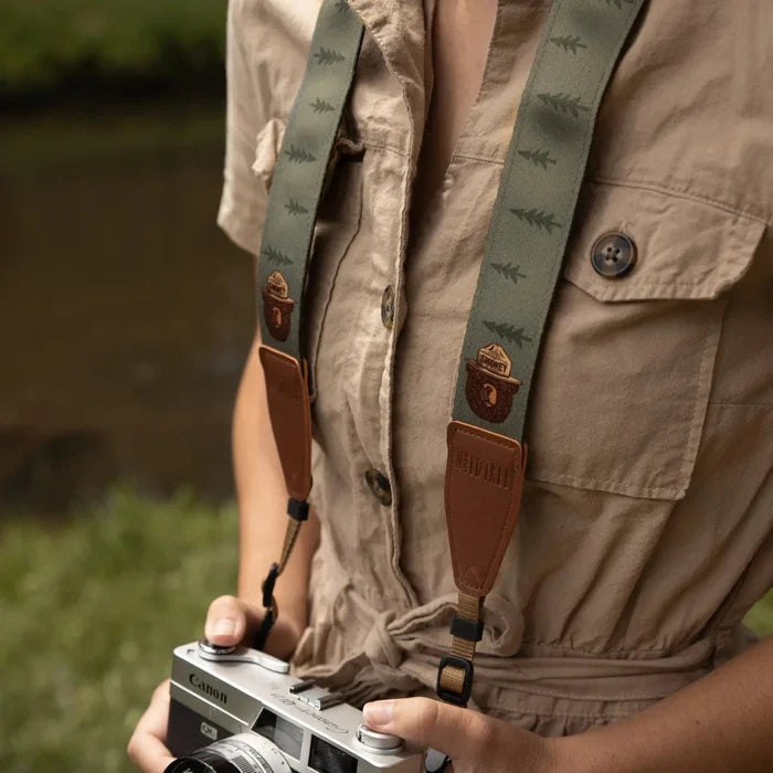 Person in tan jumpsuit holds camera with Wildtree Smokey Bear strap by grassy water, outdoor photography gear scene.