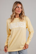 A woman wearing a pale yellow long-sleeve t-shirt with 'DAHLONEGA GEORGIA' printed in white across the chest, paired with light blue jeans, standing against a white brick wall.