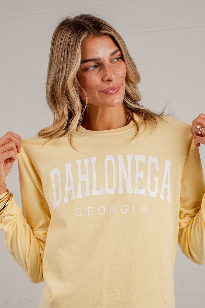 A woman wearing a pale yellow long-sleeve sweatshirt with 'DAHLONEGA GEORGIA' printed in white across the front, holding the shoulders of the sweatshirt against a light gray wall.